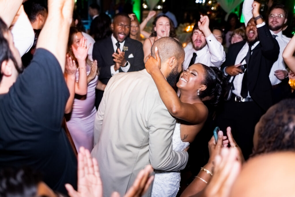 Energetic reception dance floor moment as a bride wraps her arms around her groom in the middle of a crowded celebration. Guests around them cheer, clap, and raise their hands while colorful reception lighting fills the room. The bride smiles with her eyes closed as they dance closely together, capturing the emotional reception coverage couples often prioritize when researching wedding photographer cost.