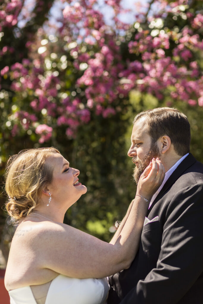 Bride gently cups the groom’s face and smiles up at him beneath blooming pink trees, a quiet and intimate portrait captured by a Central PA wedding photographer.