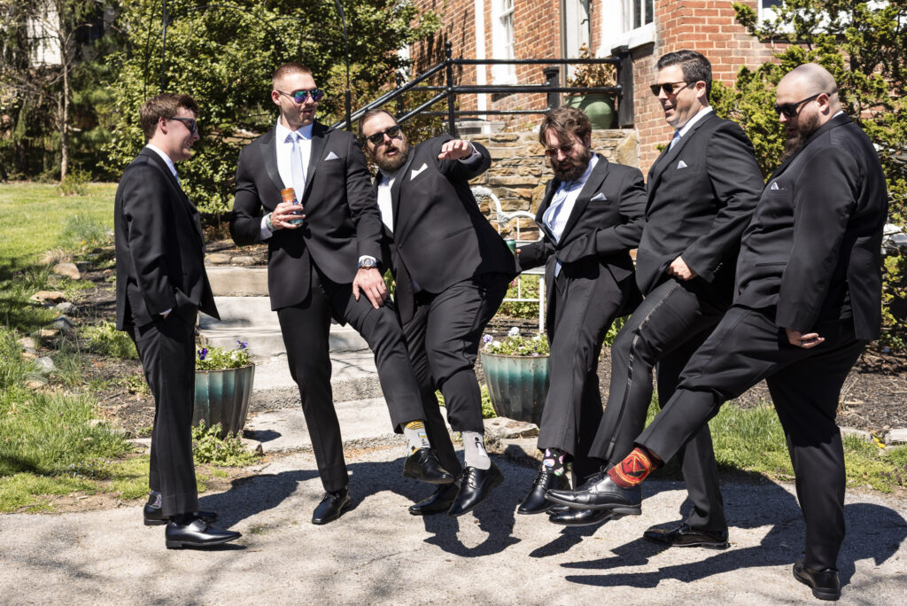 Groomsmen in black suits laugh and kick their legs in unison outside a brick building, showing off colorful socks during a playful moment at this Heritage Hills Golf Resort wedding.