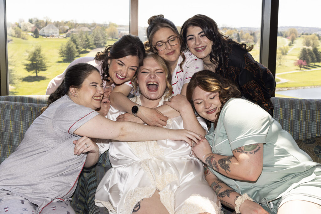 Bride sits on a couch surrounded by her bridesmaids in mis-matching pajamas, all hugging her tightly and laughing together in a joyful getting-ready moment photographed by a Central PA wedding photographer.