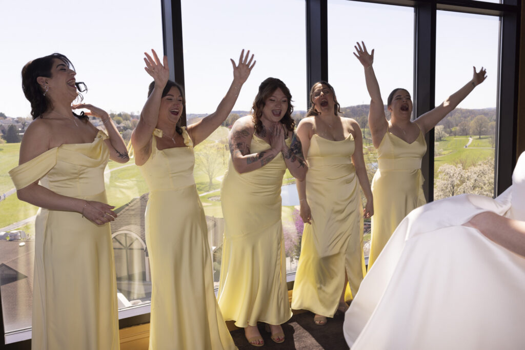 Bridesmaids in pale yellow dresses cheer with their hands raised, reacting joyfully as they see the bride for the first time in a sunlit room overlooking the grounds of this Heritage Hills Golf Resort wedding.