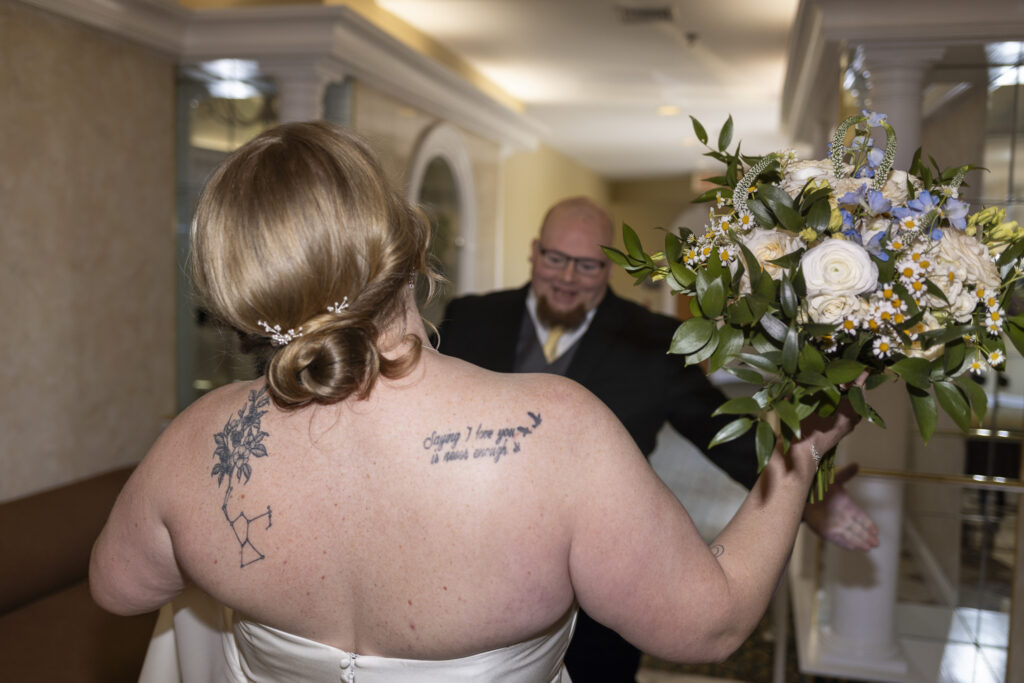 Bride holds a bouquet of white roses, blue flowers, and greenery as she approaches someone with open arms in a hallway, capturing an emotional moment during this Heritage Hills Golf Resort wedding.