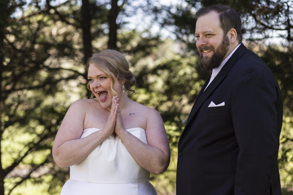 Bride reacts with excitement during the first look, hands clasped near her face and mouth open in a joyful expression as the groom smiles beside her under shaded trees.