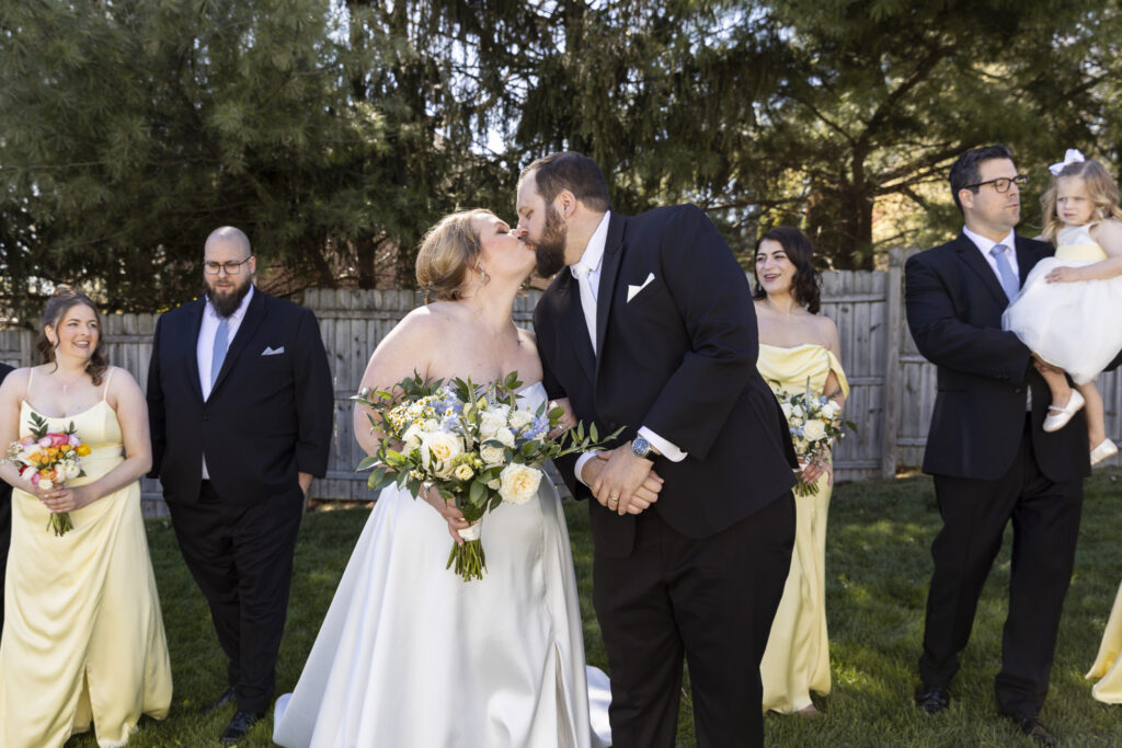 Bride and groom share a kiss while walking with their wedding party, bouquets in hand, as others smile and interact behind them in a candid moment documented by a Central PA wedding photographer.