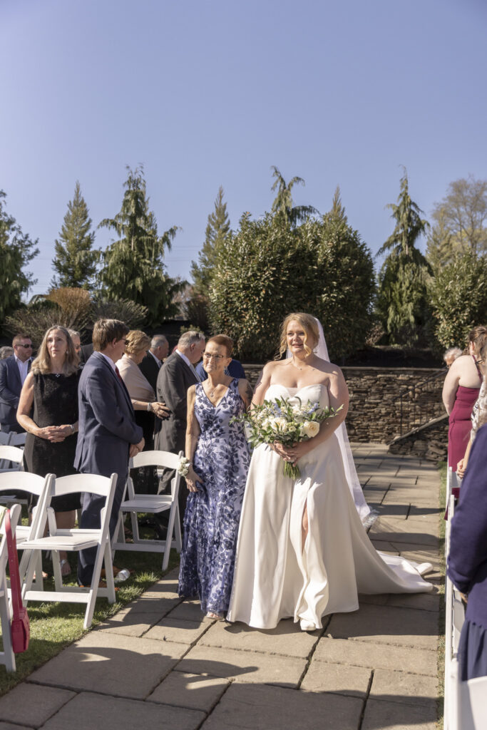 Bride walks down the aisle with an older woman, holding a bouquet of white and blue flowers, as guests stand and watch during the outdoor ceremony at this Heritage Hills Golf Resort wedding.
