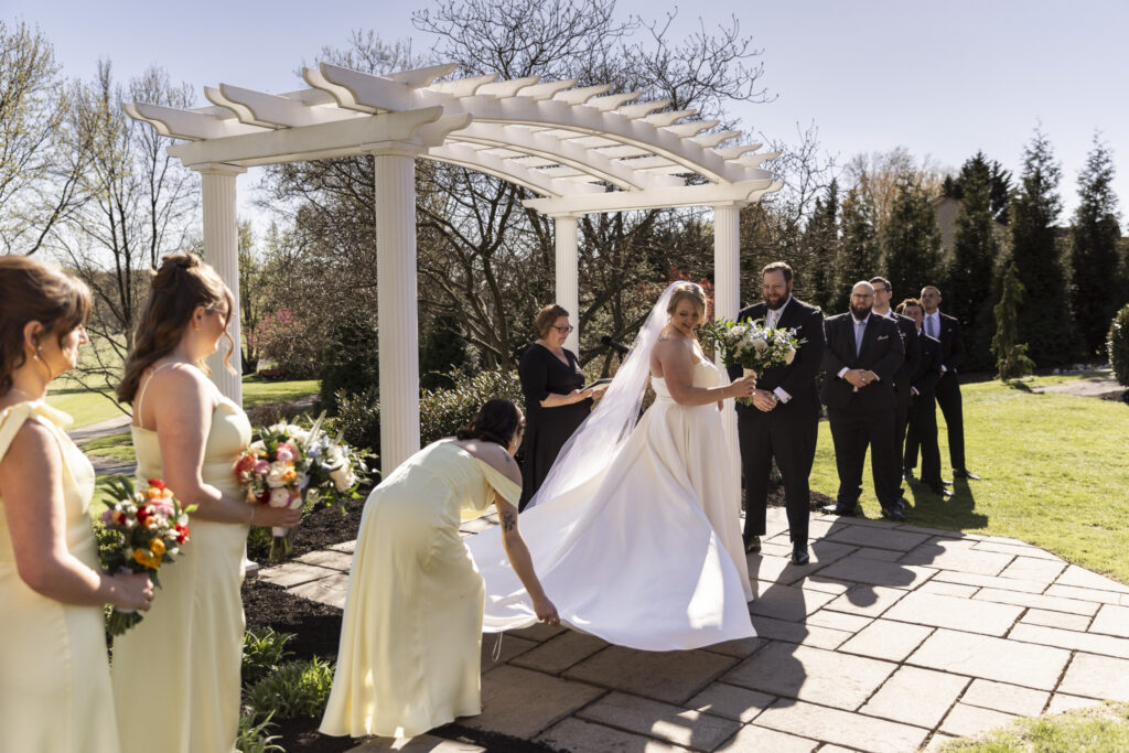 Bride adjusts her gown as a bridesmaid in a pale yellow dress fluffs the train beneath a white pergola, while the wedding party looks on during the ceremony at this Heritage Hills Golf Resort wedding.