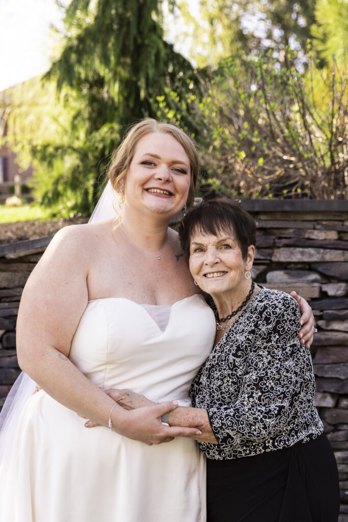 Bride embraces an elderly family member, both smiling warmly at the camera, standing in front of a stone wall with soft greenery and sunlight in the background.