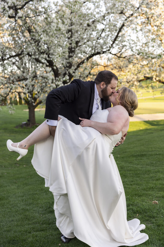 Groom lifts the bride into a dip and kisses her beneath a blooming tree during portraits at their Heritage Hills Golf Resort wedding, her dress flowing as they stand on a lush green lawn.