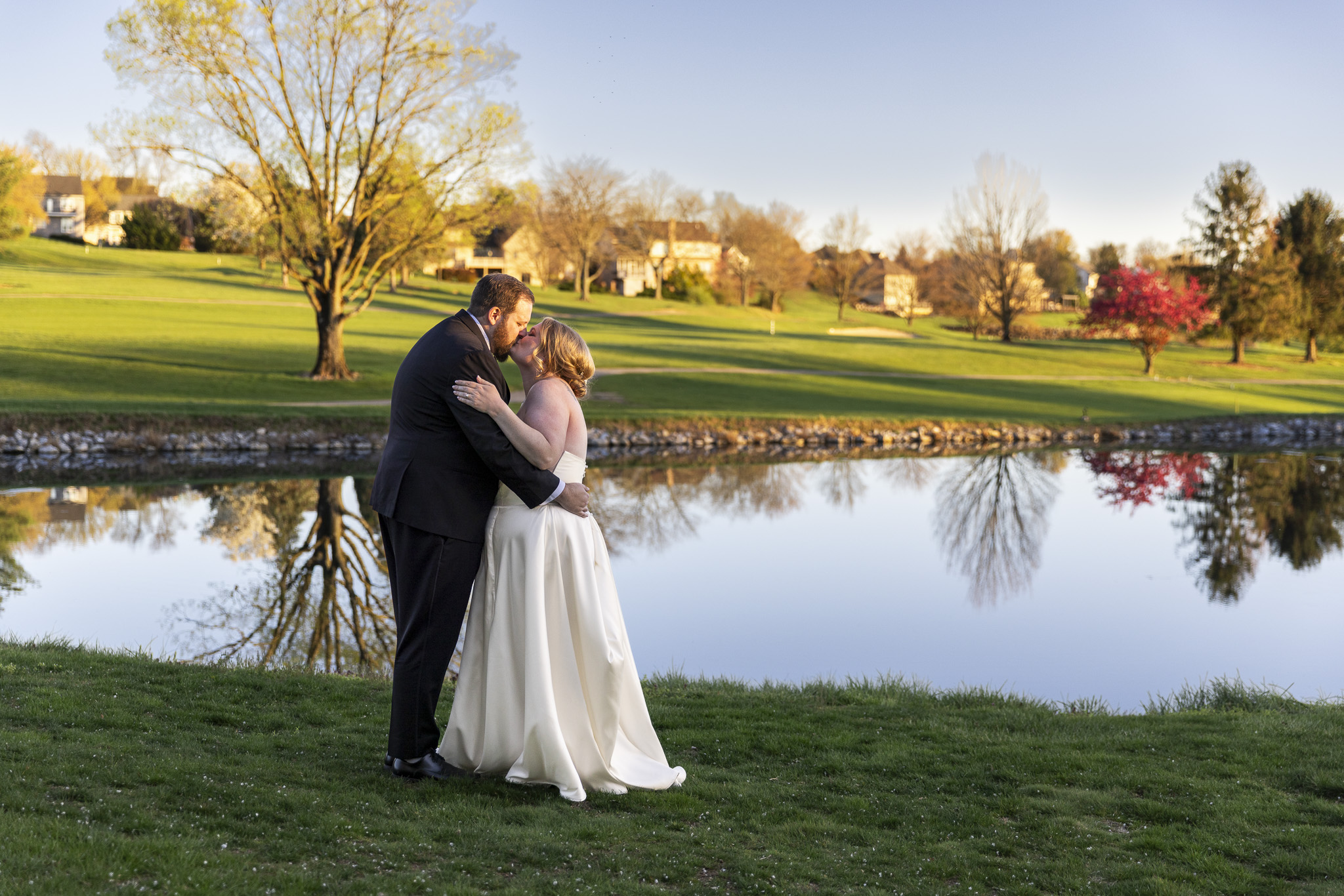 Bride and groom embrace and kiss beside a reflective pond during golden hour portraits at their Heritage Hills Golf Resort wedding, with trees and houses mirrored in the still water behind them.