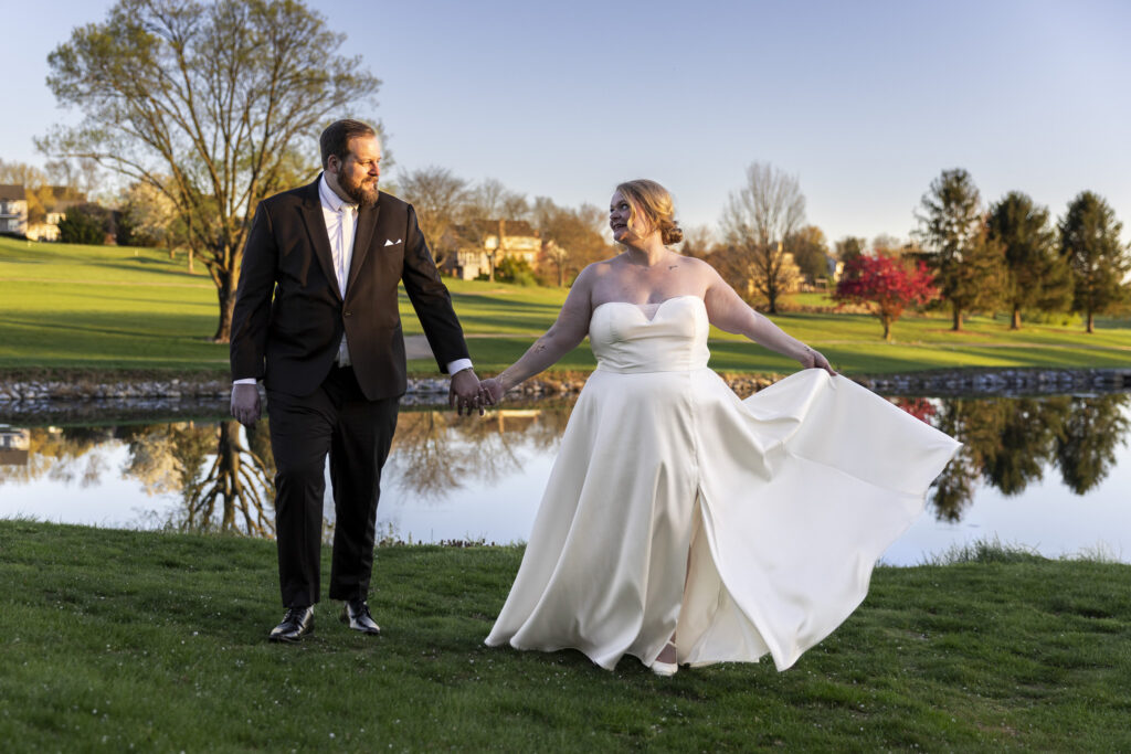 Bride and groom walk hand in hand beside a reflective pond at golden hour, her dress flowing in the breeze during sunset portraits at their Heritage Hills Golf Resort wedding.