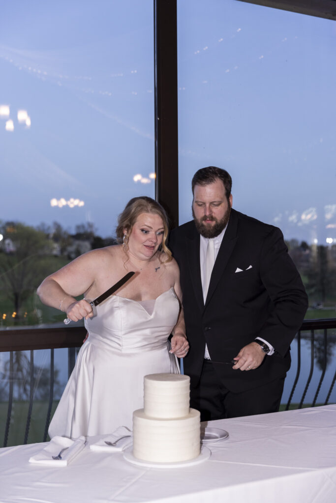 Bride and groom prepare to cut a simple white tiered wedding cake together, both leaning in with focused expressions as they prep for the first slice at their Heritage Hills Golf Resort wedding reception.