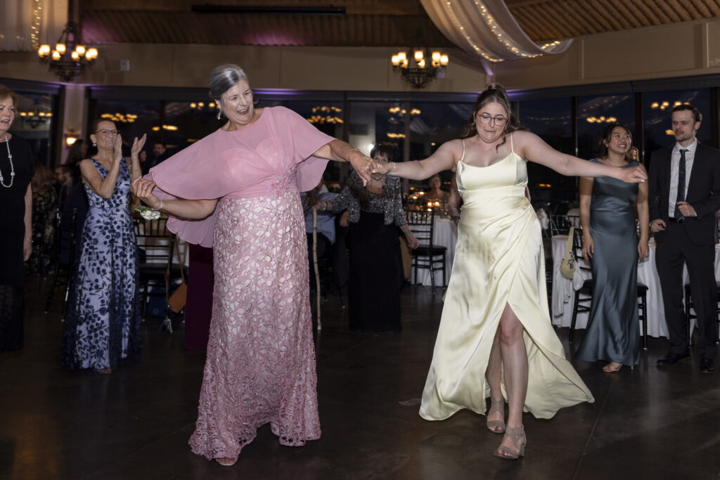 Two guests, one in a pink lace dress and the other in a pale yellow gown, hold hands and dance energetically on the reception dance floor at this lively Heritage Hills Golf Resort wedding.