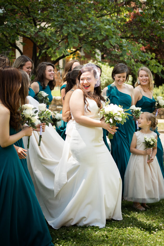A bride in a sleek white gown smiles over her shoulder while surrounded by bridesmaids in teal dresses and a flower girl holding matching bouquets. The group shares a joyful candid moment on a sunny lawn, illustrating the natural interactions that happen when couples leave enough time after asking how long do bridal party photos take for their wedding timeline.