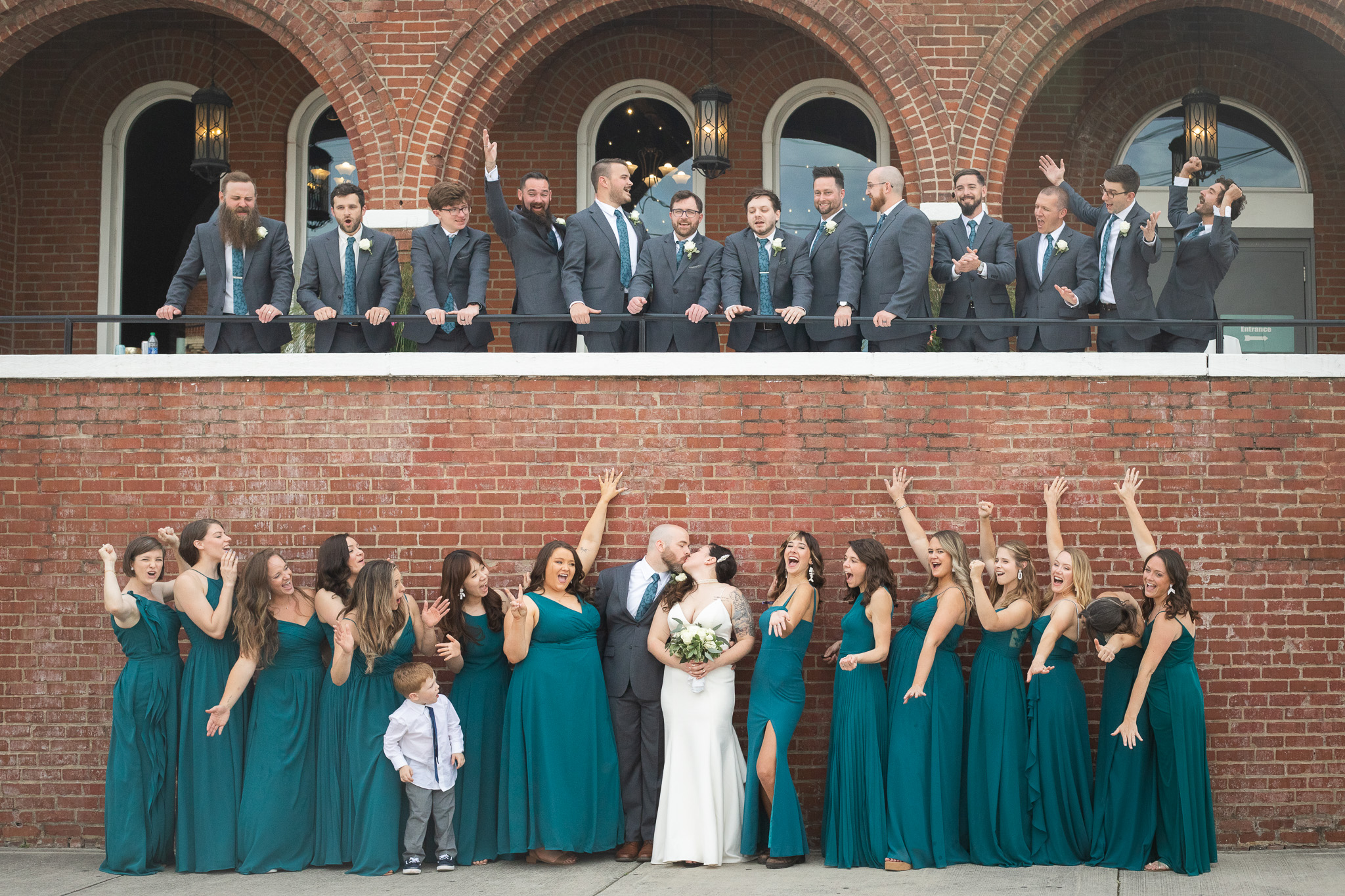 A large wedding party is split between two levels of a brick building, with groomsmen cheering from an upper balcony while the bride and groom kiss below. Bridesmaids react dramatically around them, and a young ring bearer stands near the couple looking up at the excitement.