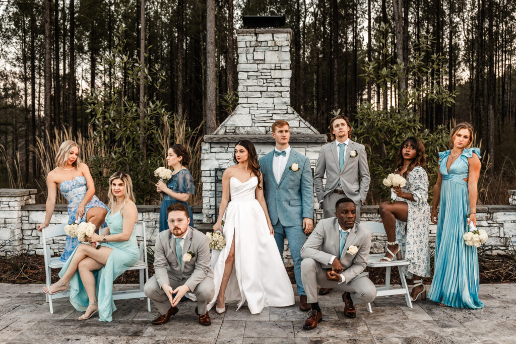A mixed wedding party poses around an outdoor stone fireplace at dusk. Some members sit while others stand confidently, creating an editorial-inspired portrait that demonstrates the variety possible during wedding party photos.