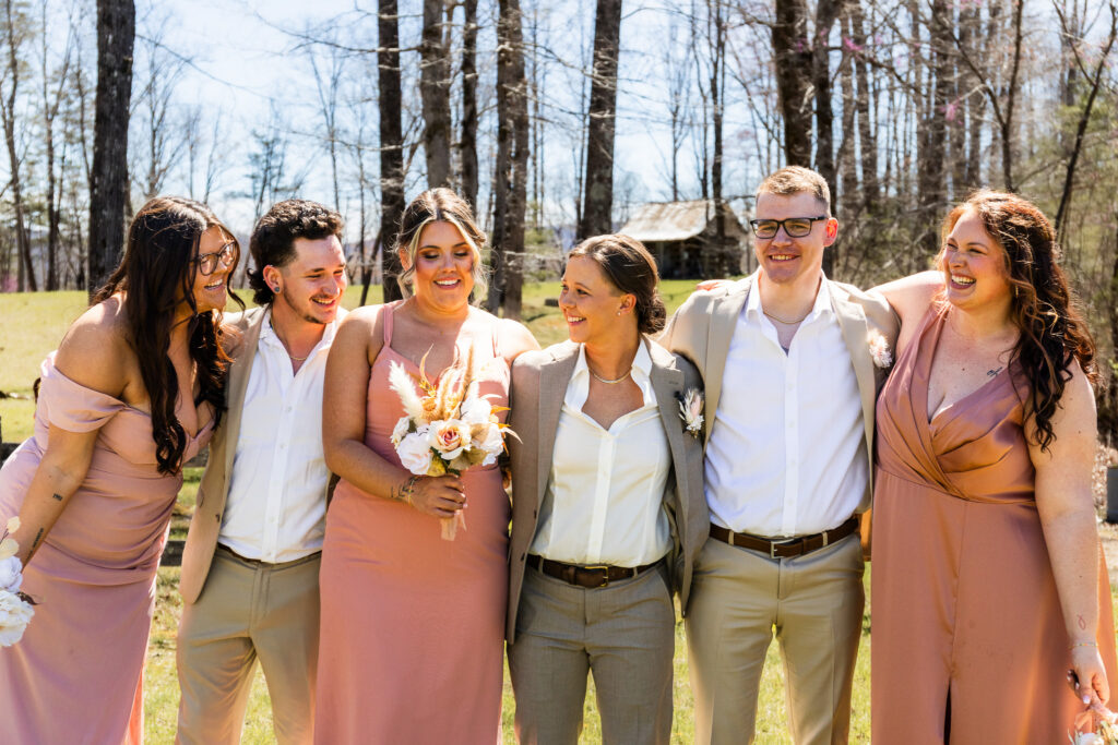 A bride in a tan suit stands arm-in-arm with five members of her wedding party, all smiling and laughing together in bright afternoon sunlight. The group forms a tight semicircle around her while she holds a bouquet of blush and cream flowers.