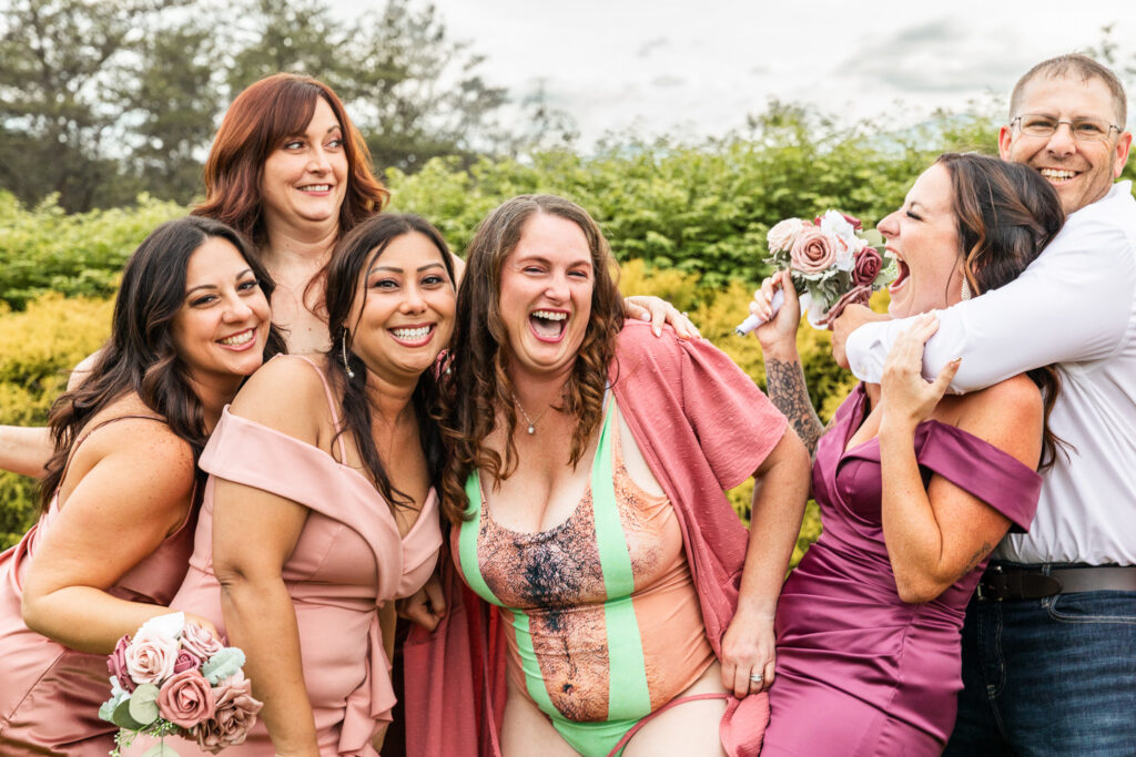 Five members of a wedding party crowd together laughing while one member of the wedding party has revealed a joke bathing suit she's wearing and another person playfully chokes a bridesmaid from behind. The candid reactions and overlapping expressions demonstrate how long do bridal party photos take can be influenced by the spontaneous moments that happen between poses.