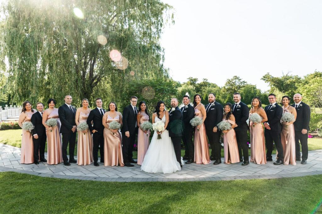 A large wedding party stands in a formal semicircle around the couple in a manicured garden. Bridesmaids wear champagne satin dresses, groomsmen wear black tuxedos, and everyone faces the camera beneath a sweeping willow tree.