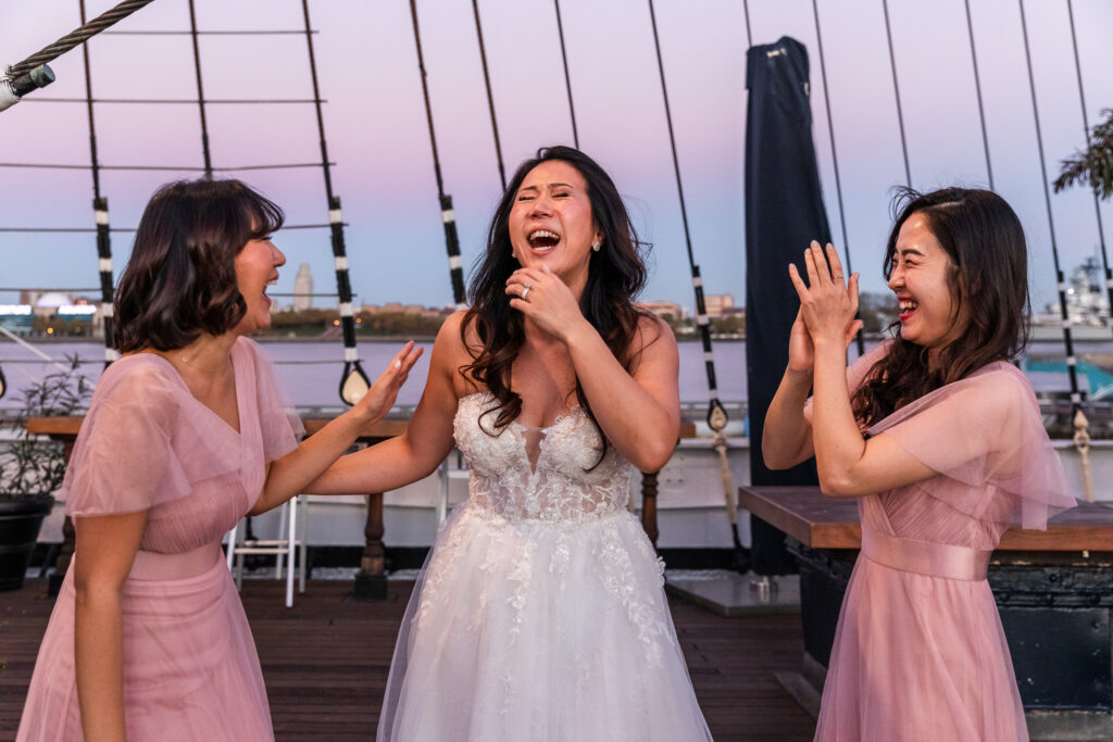 A bride in a detailed lace wedding gown laughs with two bridesmaids dressed in blush pink while standing on the deck of a tall ship at dusk. The harbor and fading evening sky create a dramatic backdrop as the three women share a genuine moment of celebration.