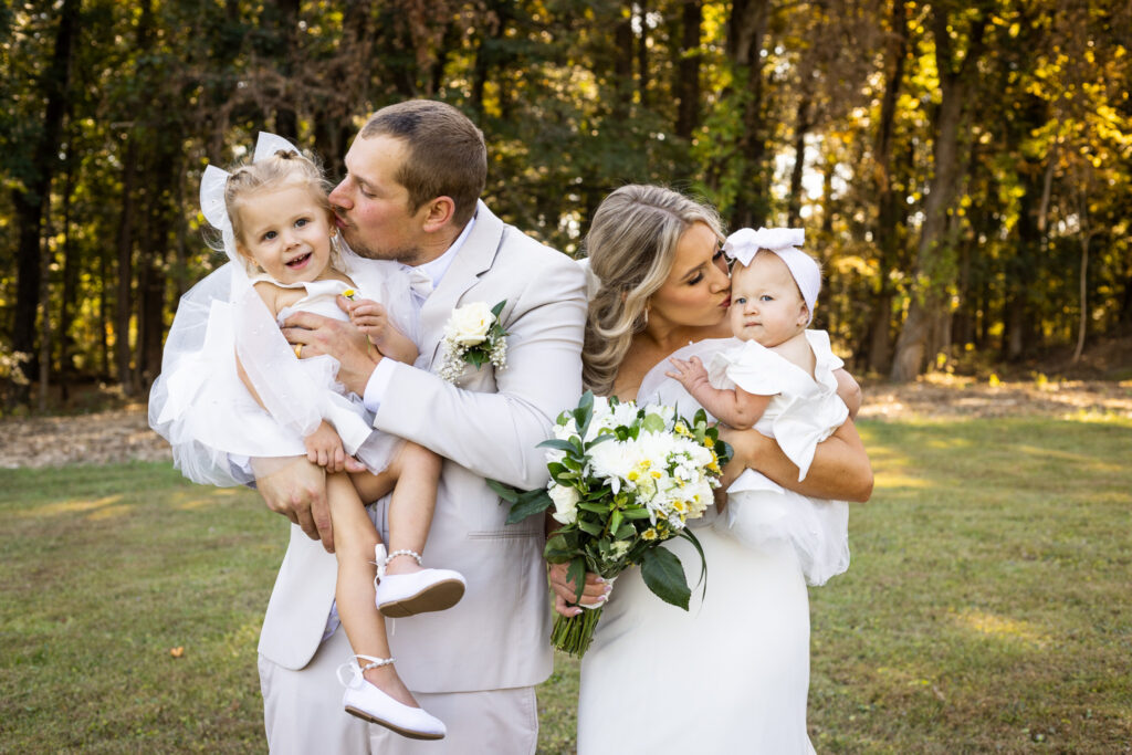 A bride and groom hold two young flower girls in their arms during portraits in a grassy field bordered by tall trees. The parents kiss each child on the cheek while the little girls look in different directions, capturing the sweet unpredictability of wedding-day family photos.