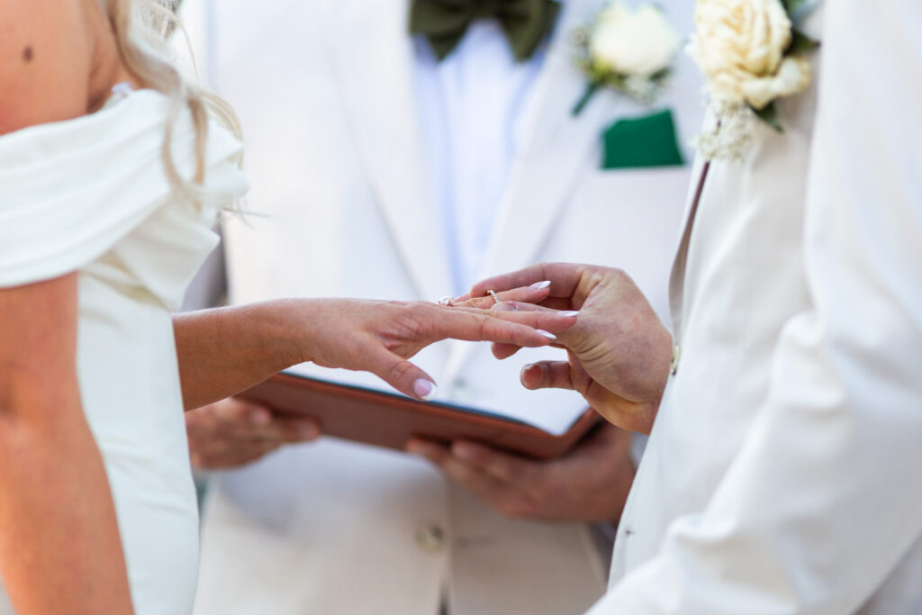 Close-up wedding ceremony photograph of a groom gently sliding a wedding ring onto the bride’s finger during their vows. The bride wears an off-the-shoulder white wedding gown while the officiant stands softly blurred in the background holding an open ceremony book. The intimate framing focuses on the emotional ring exchange and delicate details like manicured nails, wedding bands, and formal white attire.