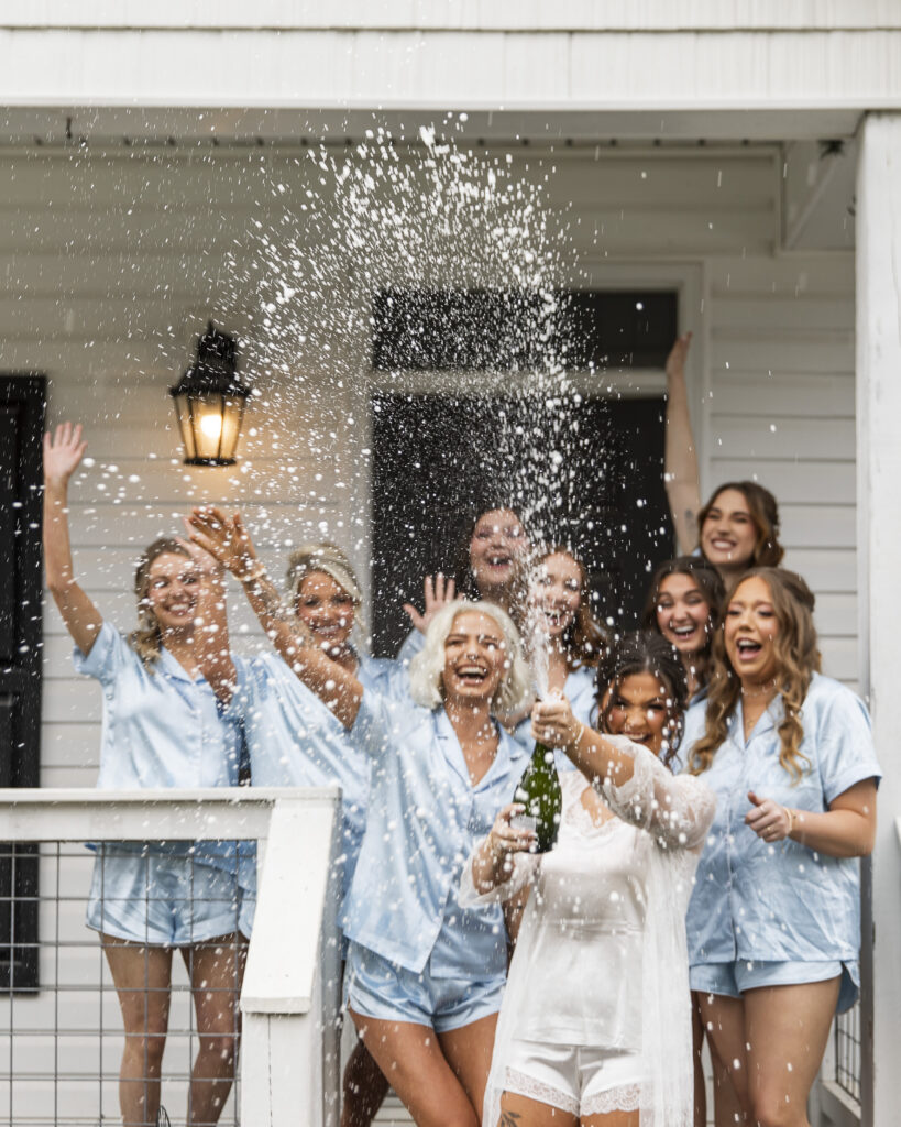 Bride and bridesmaids celebrate on the porch of a white wedding venue while popping a champagne bottle during getting ready. The bride wears white satin bridal pajamas while her bridesmaids wear matching light blue pajama sets, laughing and throwing their hands in the air as champagne sprays dramatically across the frame. The energetic candid moment and storytelling coverage highlight the kind of authentic wedding morning memories couples often value