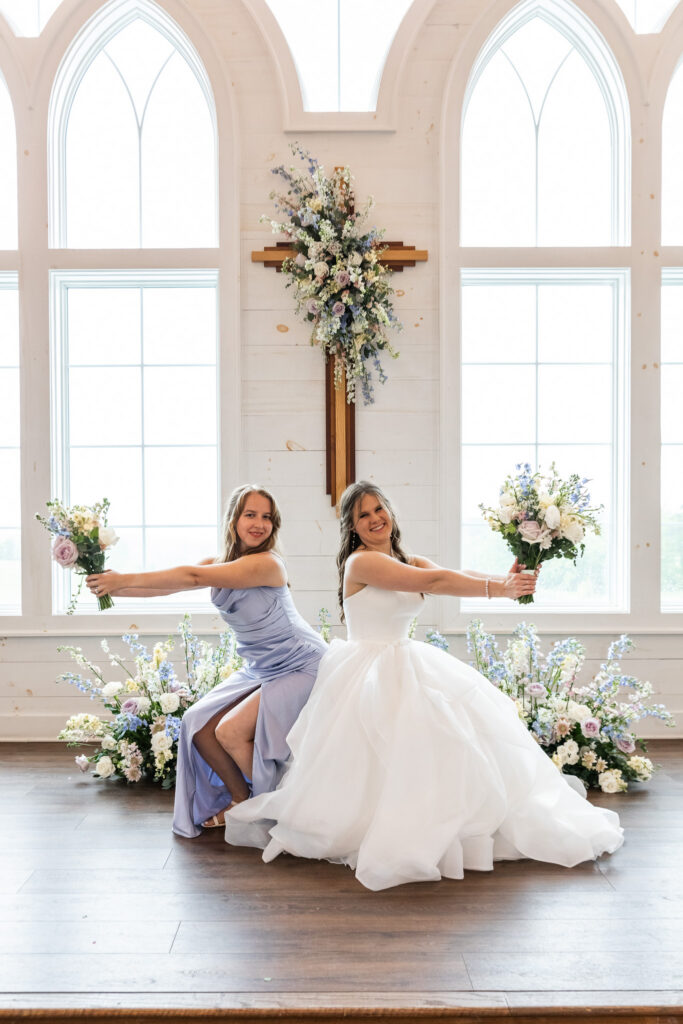 A bride and her bridesmaid sit back-to-back beneath a floral-covered cross inside a bright chapel. The bride’s white gown billows around her while both women extend their bouquets outward in a synchronized pose framed by tall arched windows and pastel floral arrangements.