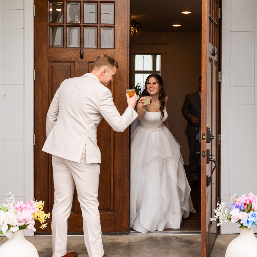 Bride and groom share a celebratory toast in the doorway of their wedding venue just before the reception begins. The groom, dressed in a light tan suit, reaches his cocktail toward the bride as she smiles from inside the doorway in a flowing layered wedding gown. Large wooden doors frame the scene while pastel floral arrangements sit on either side of the entrance, capturing the playful in-between moments couples experience on their wedding day