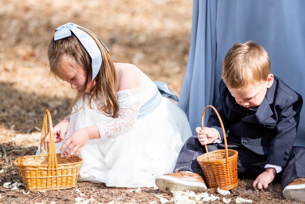 A flower girl in a white lace dress with a pale blue sash and oversized bow sits on the ground beside a ring bearer in a navy suit. Both are focused on their wicker baskets, surrounded by scattered flower petals during an outdoor wedding ceremony.