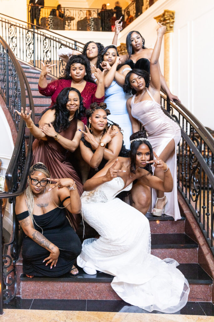 A bride crouches on an elegant staircase surrounded by bridesmaids striking playful poses and hand gestures. The group fills the staircase from top to bottom, creating an energetic portrait full of personality and movement.