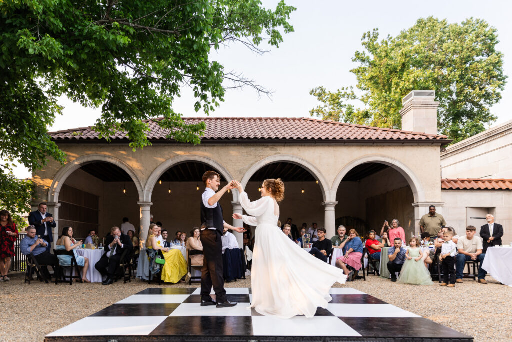 This image captures a joyful movement and documentary-style reception coverage reflect the kind of emotional storytelling couples often look for when researching wedding photographer cost. Bride twirls across a black and white checkered dance floor during an outdoor wedding reception while guests watch and smile from surrounding tables beneath stone archways and string lights. The groom reaches toward her hand as her long flowing sleeves and skirt move dramatically through the air in the warm evening light.