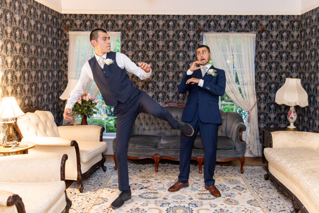 Two groomsmen in navy suits joke around in an elegant sitting room filled with antique furniture and patterned wallpaper. One dramatically kicks into the air while the other reacts in mock surprise, creating a playful wedding party portrait that demonstrates how couples wondering how long do bridal party photos take can expect time for both classic and creative photographs.