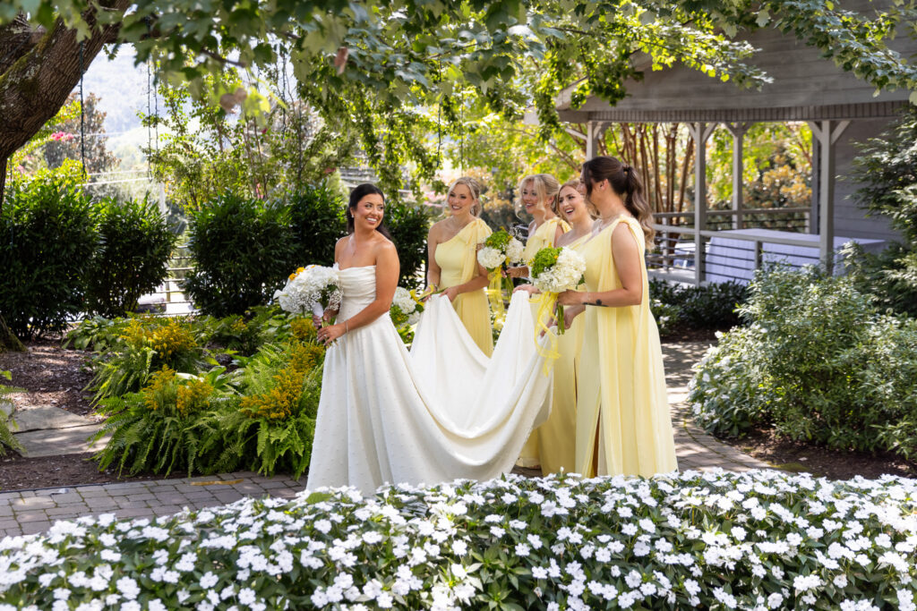 A bride in a white gown walks through a garden pathway while four bridesmaids in soft yellow dresses gather around her holding bouquets. White flowers bloom in the foreground, creating layers of depth and texture throughout the image.