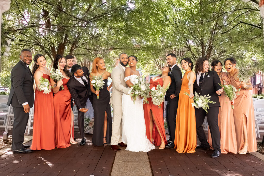 A newly married couple stands at the center of their wedding party beneath a canopy of trees. Bridesmaids in shades of rust, orange, and blush and groomsmen in black suits lean toward the couple with playful expressions while holding bouquets, creating an energetic and joyful group portrait.