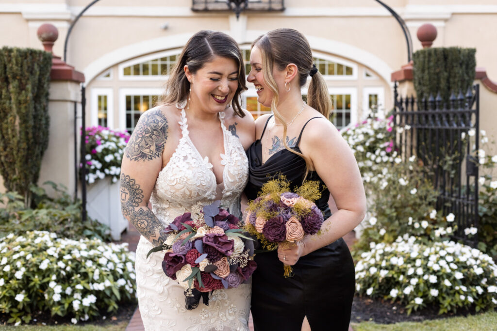 A bride and bridesmaid stand close together outside a venue surrounded by blooming white flowers. Both women hold rich purple and mauve bouquets and smile toward one another in a quiet, genuine moment.