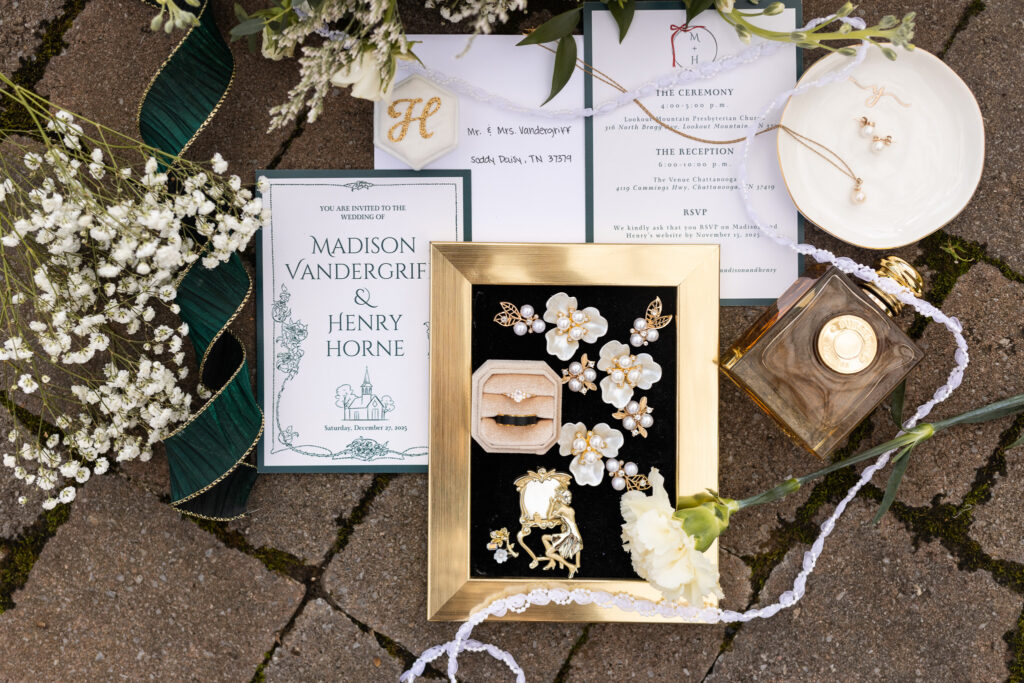 Flat lay of wedding day details arranged across stone pavers outdoors, including custom invitation suites, pearl jewelry, wedding rings, lace ribbon, perfume, floral accessories, and greenery. White flowers frame the carefully styled composition while gold accents and handwritten envelopes add an elegant, classic touch.