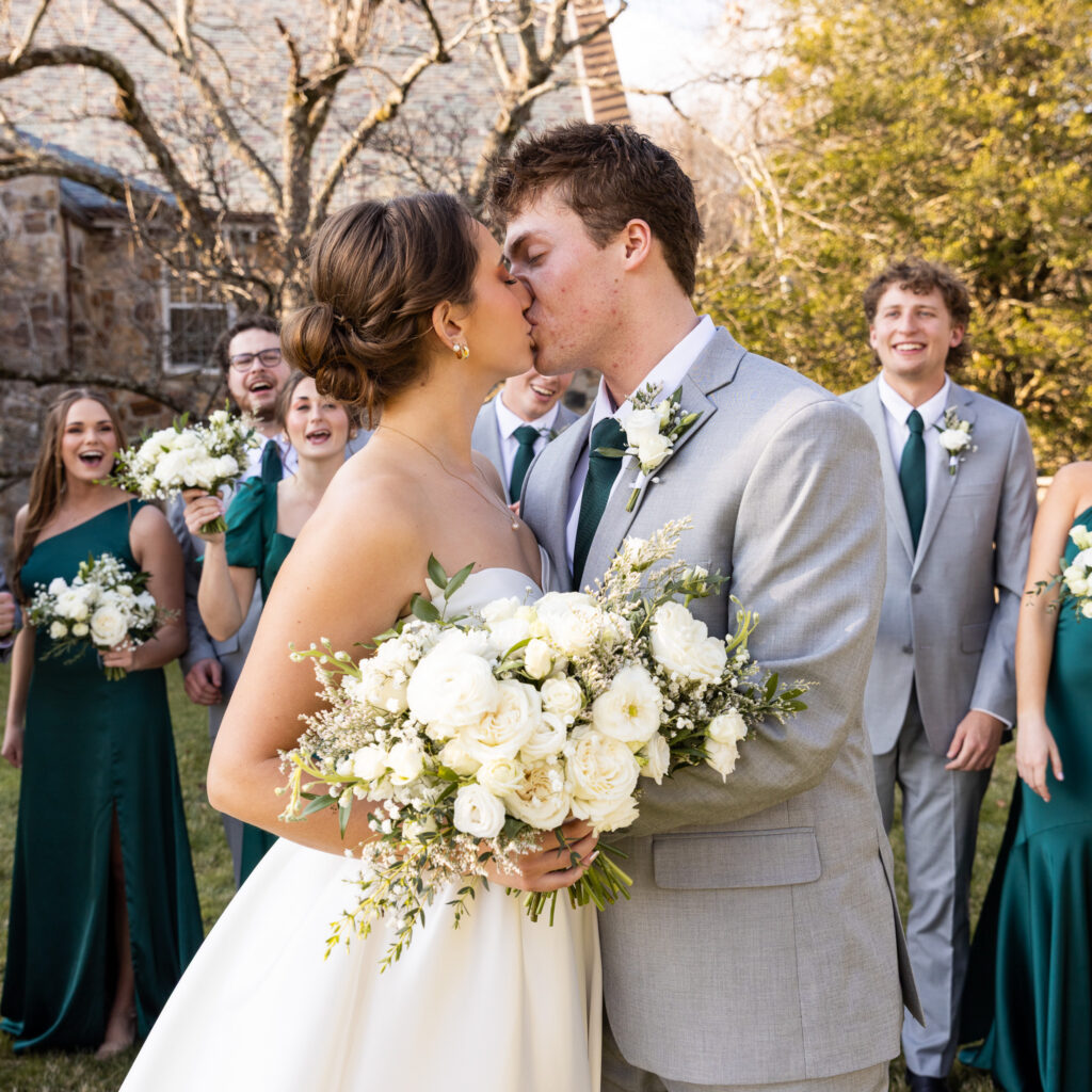 Bride and groom share a kiss outdoors immediately after their ceremony while holding a large bouquet of white roses and greenery. Bridesmaids in emerald green dresses and groomsmen in light gray suits smile and cheer around them on the lawn outside a stone church during golden afternoon light.