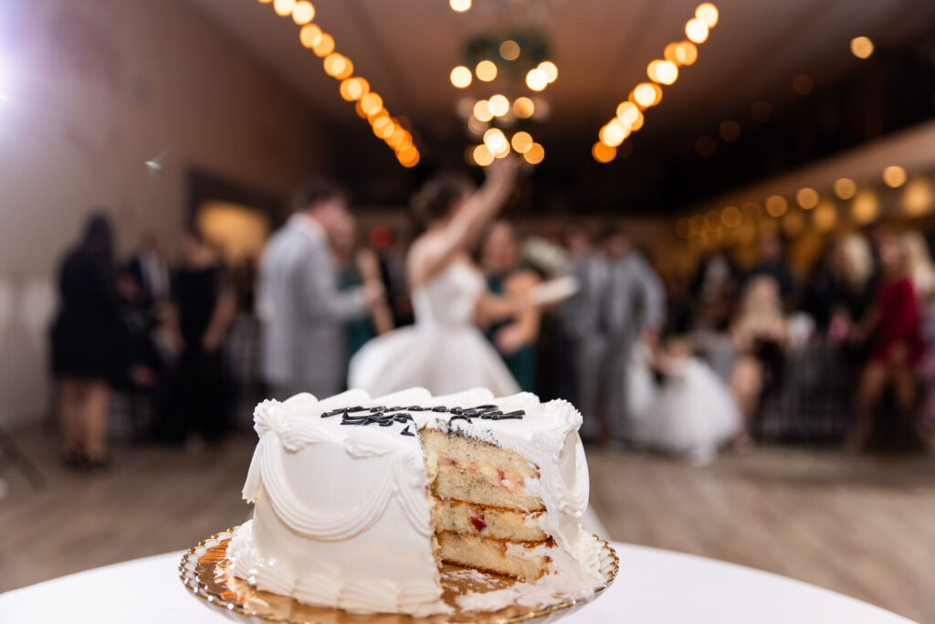 Partially cut wedding cake sits in sharp focus on a reception table while the dance floor glows softly blurred in the background. The white frosted cake reveals layers of filling inside as string lights and wedding guests create a warm reception atmosphere behind it.