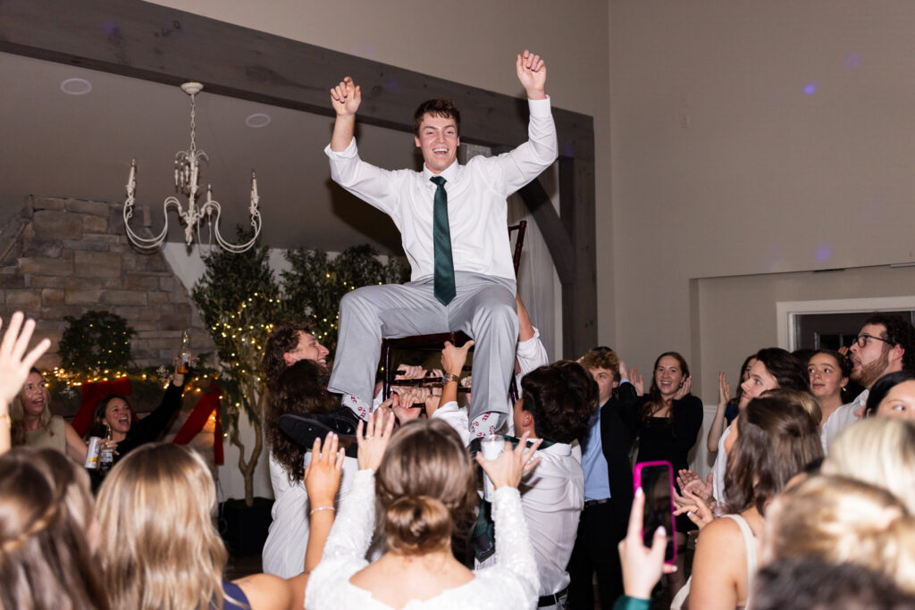 Groom is lifted high into the air on a chair during a lively wedding reception dance floor tradition while guests crowd around cheering with raised hands. He laughs with both arms in the air beneath a chandelier and warm reception lighting inside a rustic ballroom reception space.