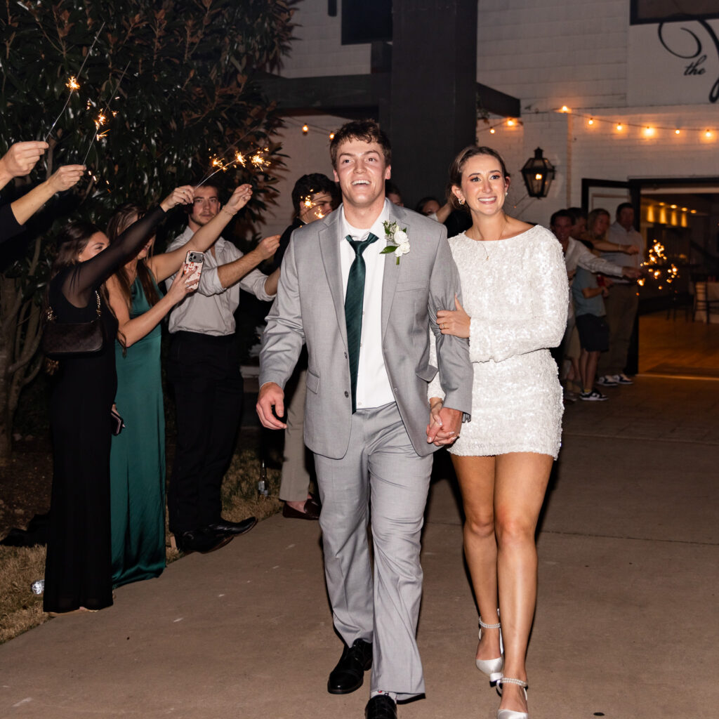 Bride and groom walk hand in hand through a nighttime sparkler sendoff outside their wedding reception venue while guests line the walkway holding glowing sparklers. The groom wears a light gray suit with a dark green tie while the bride changed into a sparkling white reception dress and silver heels for the celebration exit. Their joyful expressions and candid movement capture the kind of emotional reception coverage couples often imagine when researching wedding photographer cost.
