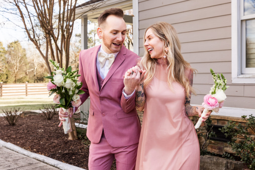 A groom in a pink suit and a member of the wedding party in a blush dress walk side-by-side outside a farmhouse venue. Both hold bouquets and smile at each other while strolling along a stone pathway, highlighting the growing popularity of mixed wedding parties.