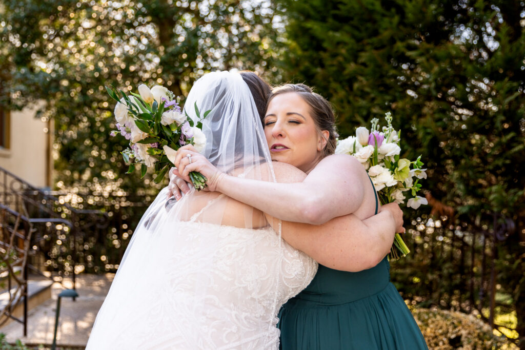 A bride in a lace wedding gown and veil embraces a bridesmaid in a deep green dress while both hold bouquets of white and lavender flowers. The emotional hug captures a meaningful friendship moment often photographed during the time couples set aside when considering how long do bridal party photos take on a wedding day.