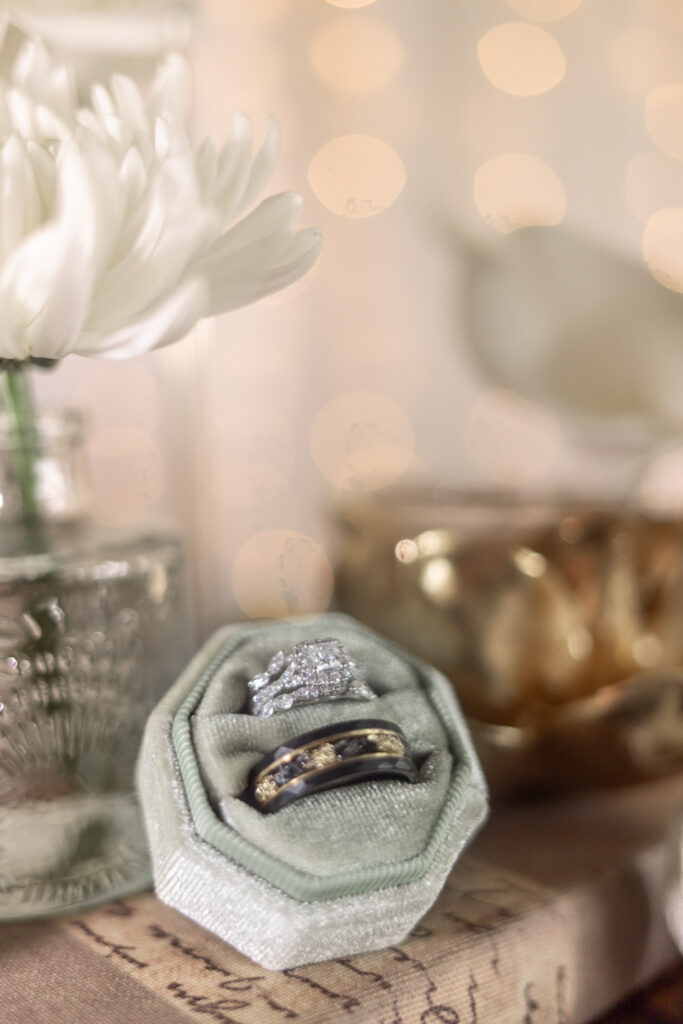 Close-up wedding detail photo of a diamond engagement ring and black-and-gold wedding band nestled inside a soft green velvet ring box, placed on top of a vintage book. In the background, a white flower in a glass vase and warm twinkle lights create a soft, romantic glow.