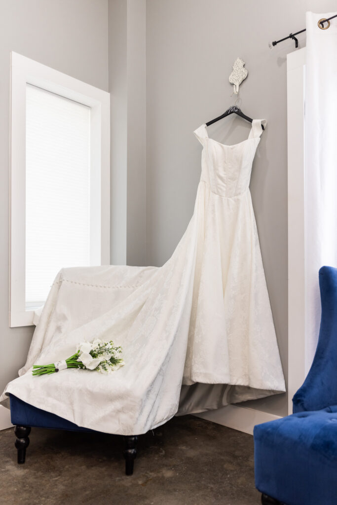 A bride’s wedding dress hangs from a wall hook in a softly lit getting-ready space, the structured bodice and full skirt draping elegantly. A bouquet of white flowers rests on a cushioned chair beneath the dress, with a blue accent chair nearby.