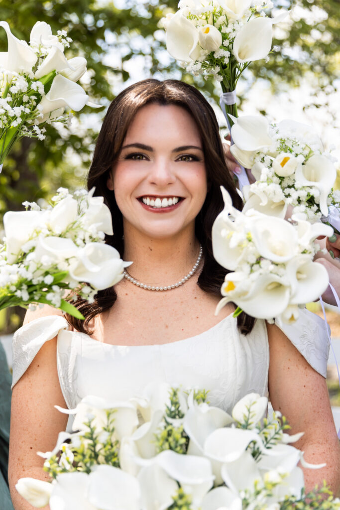 Centered in the frame during a Greenwood Oaks wedding, the bride smiles directly toward the camera while bridesmaids hold white calla lily bouquets around her face, creating a soft floral frame, with sunlight filtering through the trees behind and catching the edges of each petal and the delicate pearl necklace at her neckline.