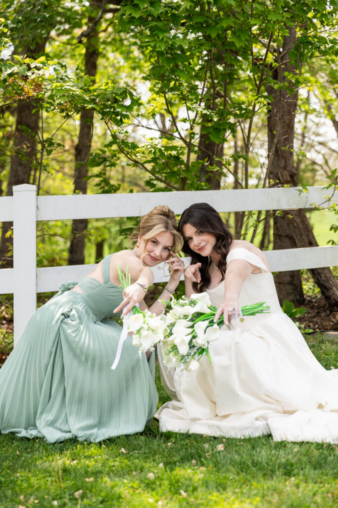 Kneeling together on soft green grass during a Greenwood Oaks wedding, the bride in an off-the-shoulder gown and a bridesmaid in a flowing sage dress lean in close beneath a white wooden fence, both reaching playfully toward the camera while holding bouquets of white calla lilies, their expressions warm and mischievous as dappled sunlight filters through the leafy trees behind them.