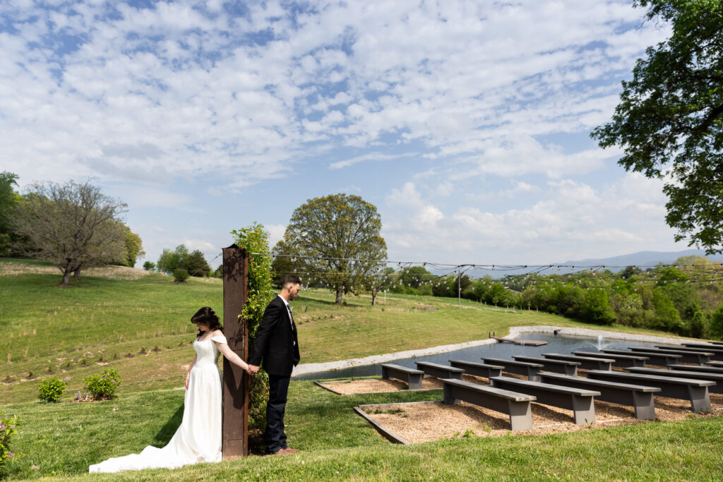 The bride and groom stand on opposite sides of a wooden post during their first touch, holding hands without seeing each other, overlooking rolling hills, a pond, and ceremony seating at a scenic Johnson City wedding venue.