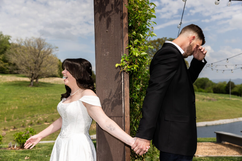 Emotional first touch moment where the bride smiles and reaches back while the groom wipes his eyes, both holding hands around a wooden post wrapped in greenery at an outdoor Johnson City wedding venue.
