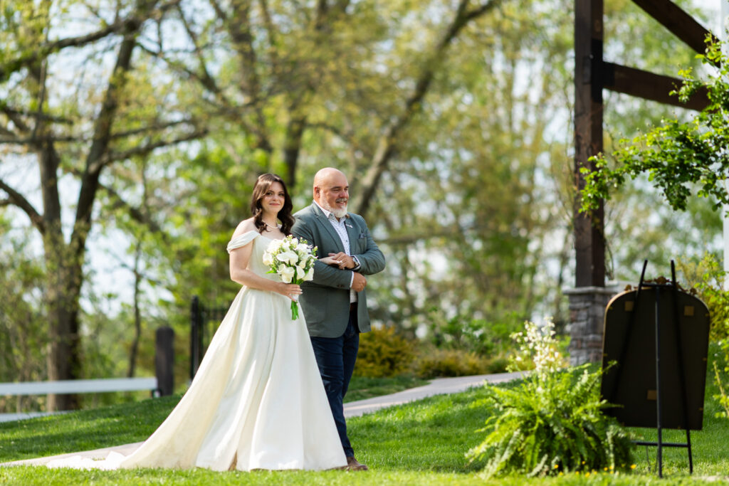Walking down the aisle at a Johnson City wedding venue, the bride is escorted arm in arm by an older man, holding a bouquet of white flowers as guests look on from either side.