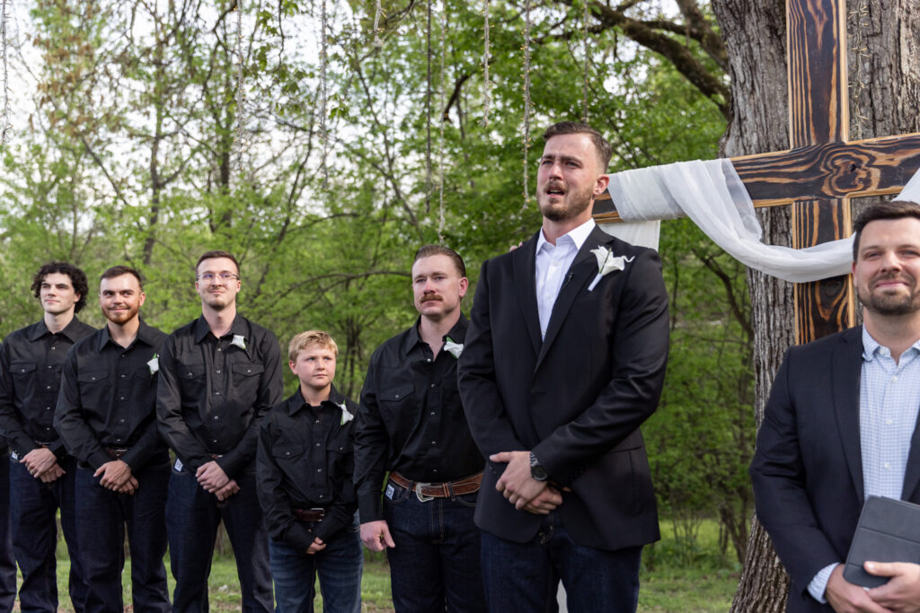 At the altar of an outdoor Johnson City wedding venue, the groom stands with his groomsmen, visibly emotional beneath a wooden cross draped in fabric and surrounded by trees.